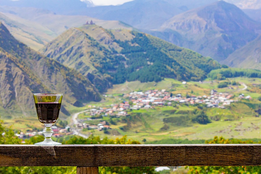 A glass of Georgian red Wine stands on a wooden railing on the background of the Georgian village and Kazbeg mountain. Near the village of Stepantsminda on the Georgian military road. Kazbegi Georgia A glass of Georgian Wine on the background of the Georgian village and Kazbeg mountain near the village of Stepantsminda on the Georgian military road. Kazbegi Georgia
